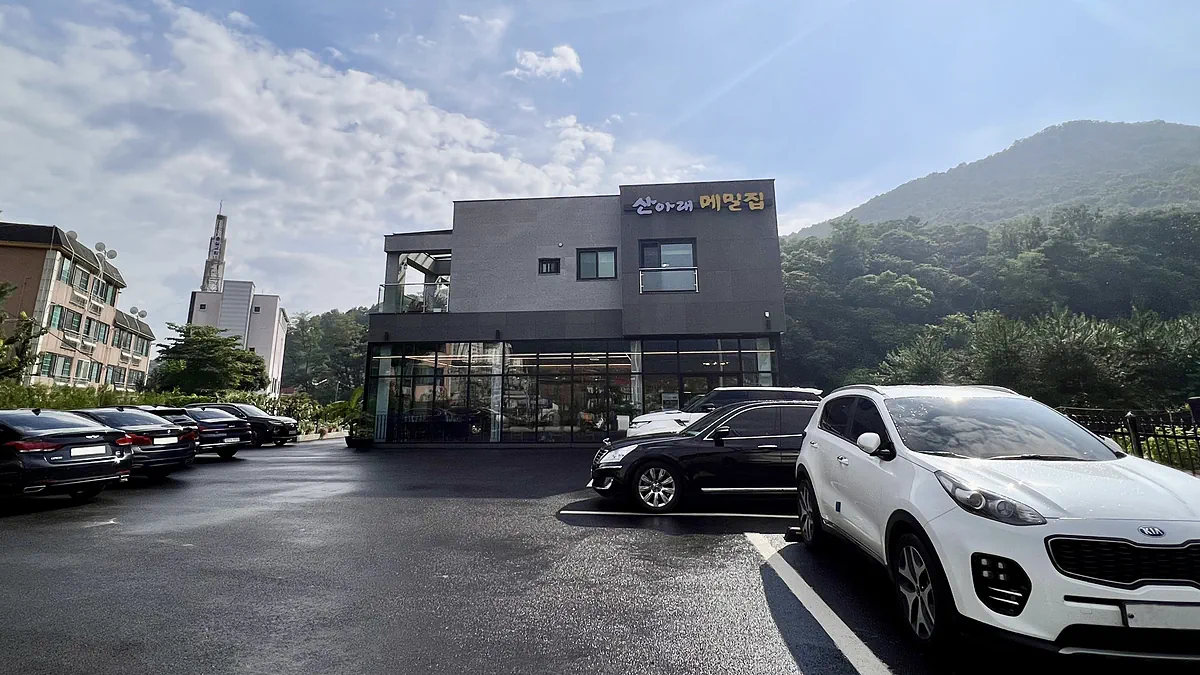 산을 등지고 선 산아래메밀집 건물. 2층 외벽에 간판이 있고 앞쪽이 주차장이다